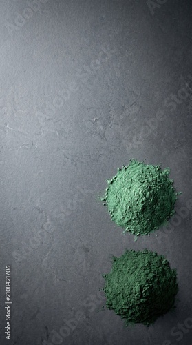 Spirulina powder sits in two small mounds on a smooth, dark stone surface. The morning light highlights the deep green color, showcasing its vibrant texture and healthful properties.