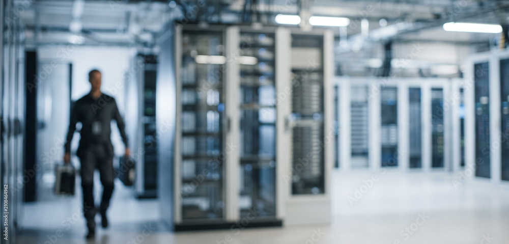 Obraz premium A blurred image of a data center featuring server racks and a person walking through. The environment is modern and technology-focused, highlighting infrastructure.