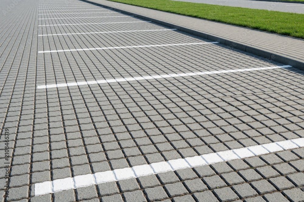 Fototapeta premium Empty parking area paved with grey interlocking stones and white lines. Green grass borders the walkway next to the car spots. Modern outdoor urban ground surface.