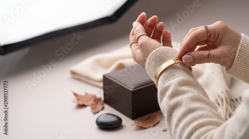 Elegant woman fastening gold cuff bracelet wearing soft beige knitted sweater in cozy setting with carefully fastening textured close person wearing
