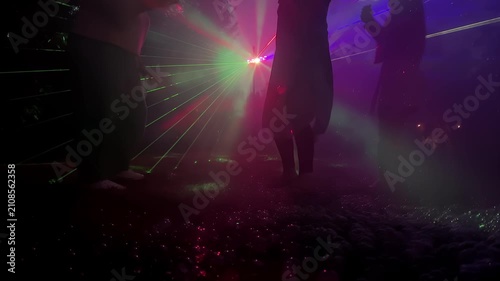 A group of friends dances together under colorful laser lights and bubbles in an outdoor setting at night, with vibrant beams illuminating the scene and creating a festive atmosphere