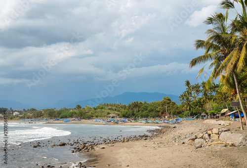 Scenic view of Medewi Beach in Bali, Indonesia, featuring traditional fishing boats, a tropical coastline, swaying coconut palms, distant mountains, and dramatic clouds.