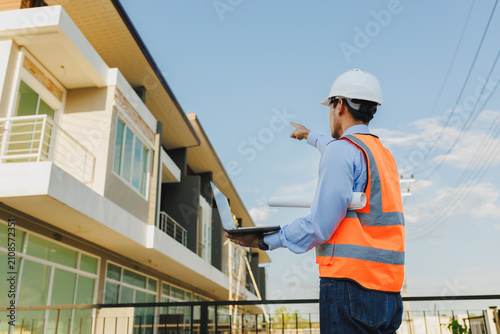 Asian engineer from behind wearing safety vest and hard hat, pointing at a townhouse while holding a laptop at a construction site.