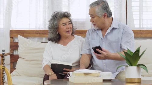 Happy Asian senior couple sitting together on couch at home man showing smartphone screen to his wife and smiling while playing phone
