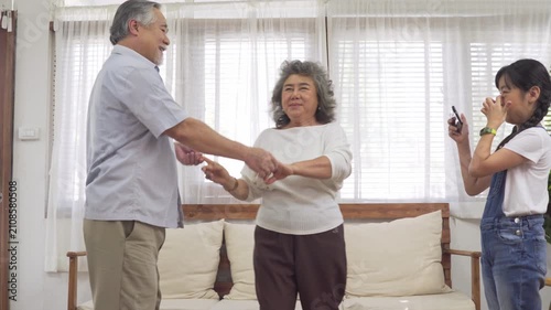 Little Asian girl taking photo with smartphone while senior grandfather and grandmother holding hands dancing together in living room at home happy family moment