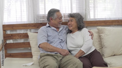 Happy senior Asian man embracing wife sitting together on couch in living room at home sharing moment of love care and support during retirement