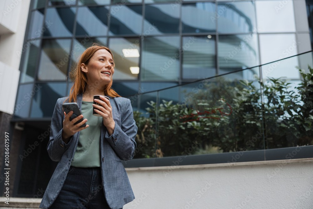 Fototapeta premium Businesswoman using her smartphone and drinking coffee outside an office building during a work break.