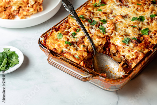 A baking dish of freshly made baked ziti hot out of the oven, with one serving.