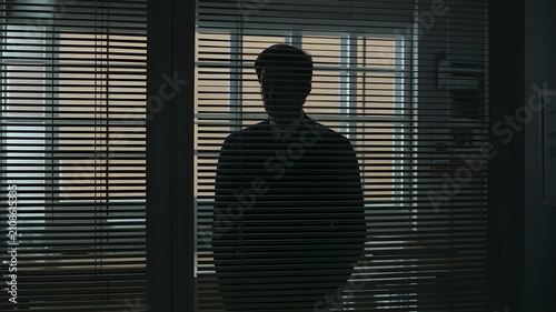 Silhouette of a man standing behind window blinds in a dark room, mysterious figure looking through horizontal slats, dramatic lighting and moody atmosphere