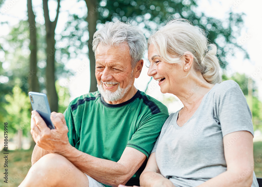 Fototapeta premium Happy active senior couple having fun using smartphone and wearing sportswear, after having an exercise sport activity outdoors