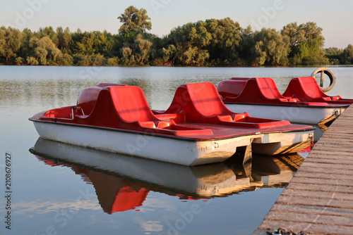 Red pedal boats sit by the water in the late afternoon sun near a tree-lined shore