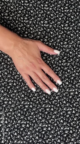 A woman's hand with a manicure gently runs over a black fabric with a small white floral pattern, demonstrating the texture of the material. Concept: fabric selection, sewing materials, tactile