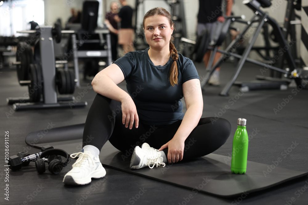 Fototapeta premium Happy overweight woman with bottle of water sitting on fitness mat in gym