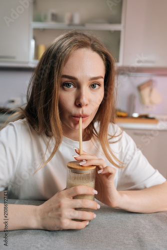 Vertical portrait of young caucasian woman enjoying healthy smoothie at home. She sips from glass with eco friendly straw, looking at camera. Soft light, shallow depth of field, wellness concept.