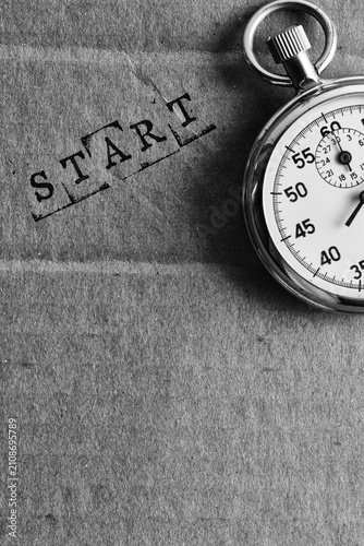 Black and white photo mechanical stopwatch next to stamped word START on paper texture.
