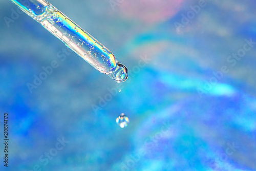 Macro shot of a glass dropper releasing a water droplet over an iridescent blue background for skincare