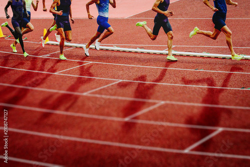 Running long distance track and field runners in motion on a red track, featuring low-angle legs, dramatic shadows, and a blurred foreground with edit space.