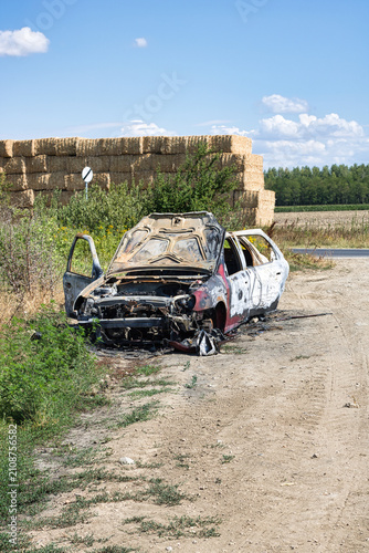 Horizontal front wide shot of a completely burned-out white car wreck abandoned on a dirt roadside.
