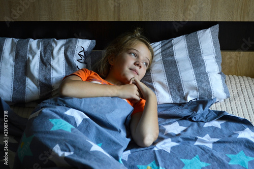 A girl in an orange T-shirt is lying on a pillow. The child is smiling and looking away. The child has woken up, is lying relaxed, and is enjoying the new day.