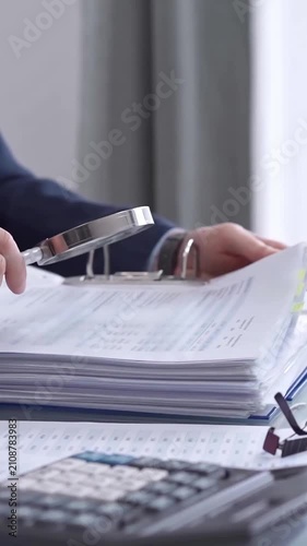 Businessman reviewing ring folder of financial documents with magnifying glass at desk in modern office. Audit and taxes in business