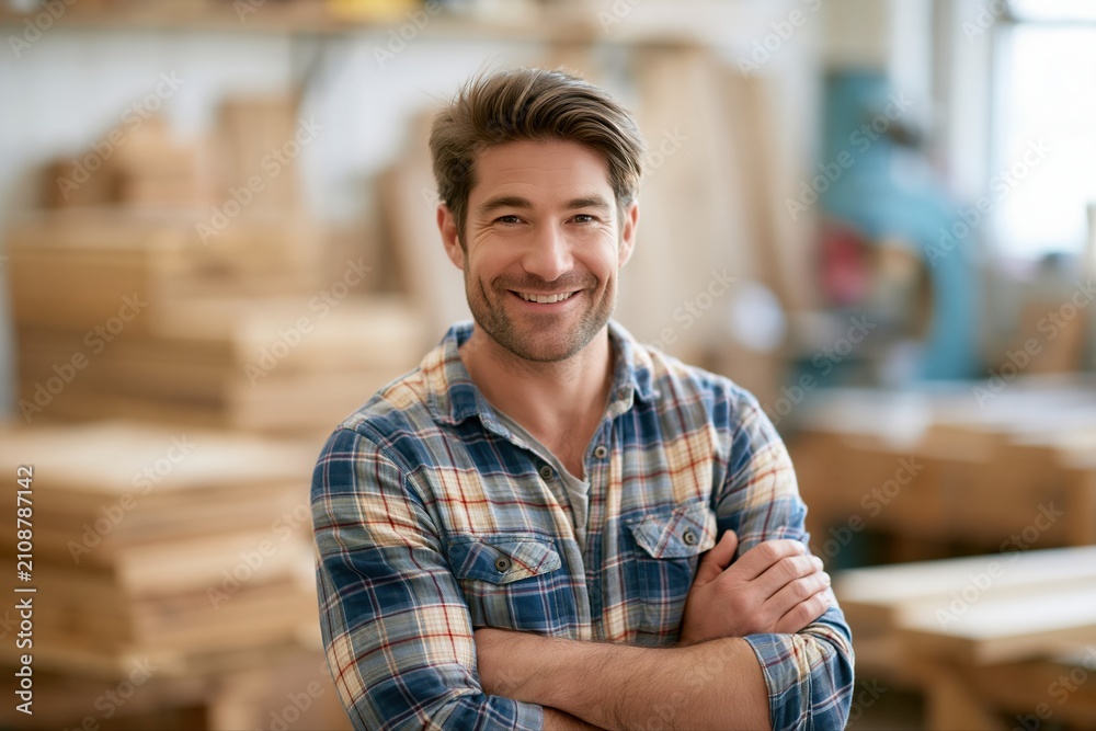 Obraz premium Smiling Caucasian Carpenter in Plaid Shirt with Arms Crossed in Wood Workshop
