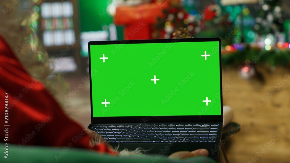 Fototapeta premium Santa Claus reads a letter and children's Christmas wish lists on a green screen laptop, using a chroma key notebook while wearing a festive red costume during the holiday season.
