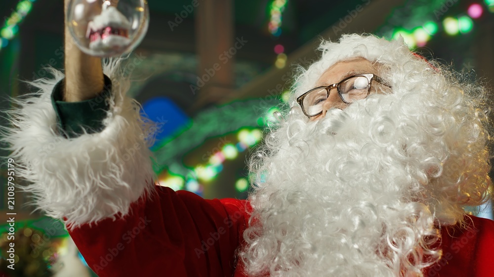 Fototapeta premium Santa Claus decorates his workshop with Christmas ornaments and a globe while preparing the festive room for photographs with visiting families.
