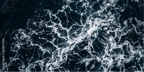 A close-up aerial view reveals the intricate patterns of dark ocean water with white foam swirling, creating a dynamic abstract texture.
