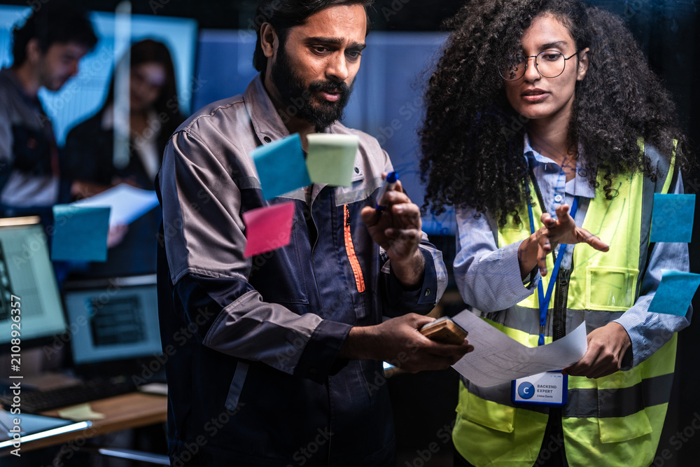 Obraz premium Operators work at a long bank of glowing monitors in a dark control room filled with streaming data at night, with rows of screens glowing in the dark behind while the city lights glow.