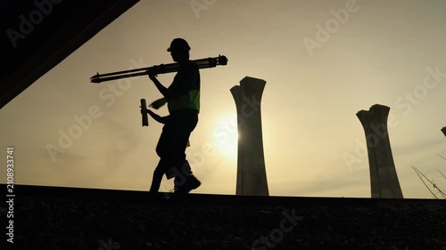 Silhouette of a survey engineer carrying a tripod across a construction site during sunset with concrete bridge columns in the background, symbolizing engineering, infrastructure, and fieldwork.
