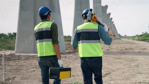 Two construction engineers walking through an infrastructure construction site carrying surveying equipment and field toolbox for engineering inspection and site measurement.
