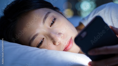 A young Asian woman is lying in bed in a dark room, illuminated by the glow of her smartphone screen as she uses it late at night.