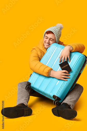 Handsome young happy man with suitcase and passport sitting on yellow backgro...