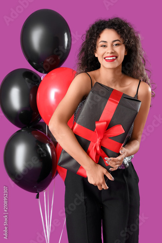 Beautiful African-American woman with gift box and balloons on purple backgro...