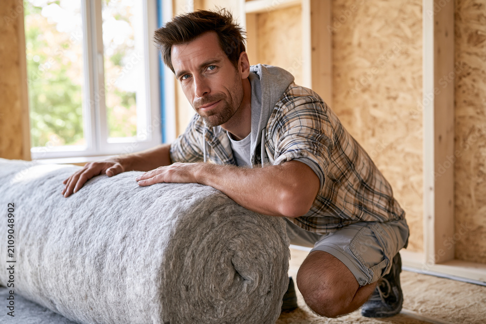 Obraz premium Man kneels beside insulation roll in unfinished room. He looks at camera with focused, determined expression. Natural light streams through window beside him