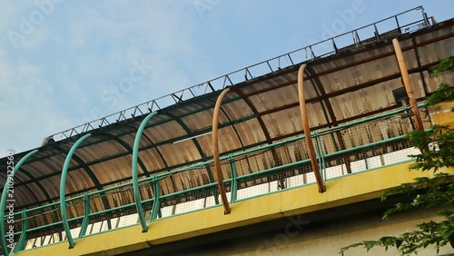 Obraz na plátně A colorful urban pedestrian overpass with a curved roof structure and green metal railings, viewed from below and framed by foliage against a clear blue sky