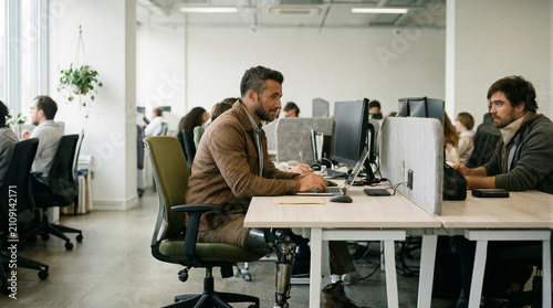 Man with prosthetic leg working on laptop at desk in open office