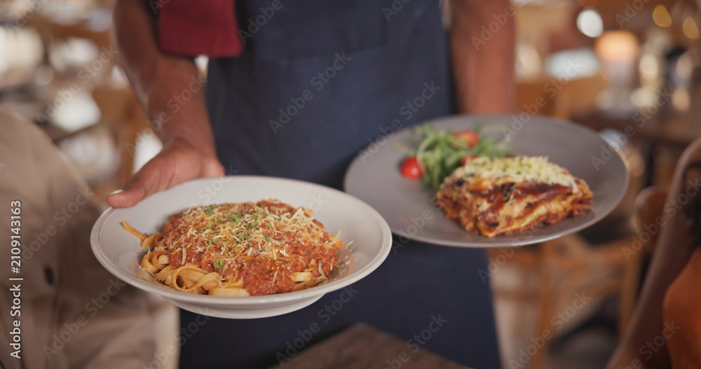 Obraz premium Hands, waiter and food at restaurant with couple, gourmet meal and fine dining for anniversary. Employee, serving and people with plate for Italian cuisine, spaghetti dish and romance for celebration