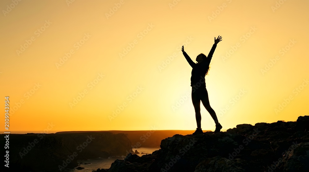 Fototapeta premium Full body silhouette of a woman hiker standing on a rocky cliff edge with arms raised toward a bright yellow sunset sky over a coastal landscape and ocean horizon.