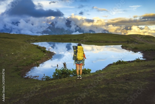 Woman hiker with yellow backpack stand by mirror-like Koruldi Lake enjoy sunset in Georgia. A peaceful mountain landscape in Svaneti with reflections, hiking, and pet-friendly adventure in Georgia Alps