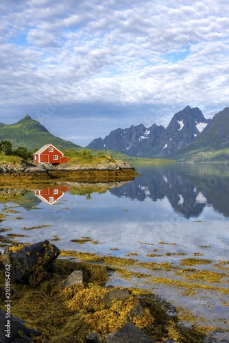 Red fishing hut on the shore, Rorbuer hut, picturesque fjord landscape at Raftsund fjord, mountains reflected in water, Vesterålen, Norway