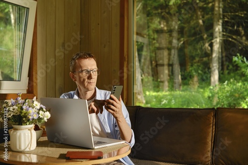 Middle-aged nomad sits at table in wooden cabin using laptop and smartphone. Sunlight shines through windows. Green trees visible outside. Scene shows focus on work amidst natural surroundings