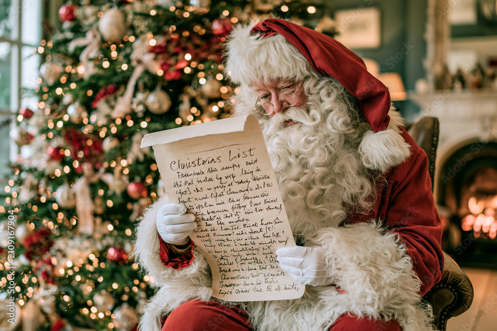 Fototapeta premium Santa Claus sits cozily by a glowing fireplace. He holds a handwritten Christmas list with gentle smile. The tree behind him sparkles with warm lights and ornaments