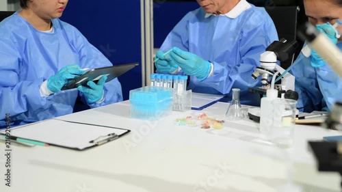 Scientific team in protective gowns collaborating in a modern lab, using a digital tablet and microscope to analyze experimental samples.