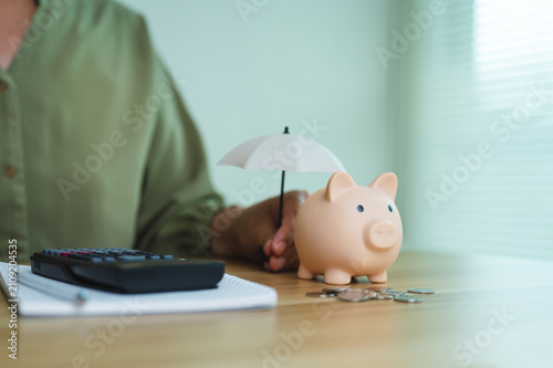 Insurance concept and financial planning, hand holding small umbrella over piggy bank on desk with calculator and coins