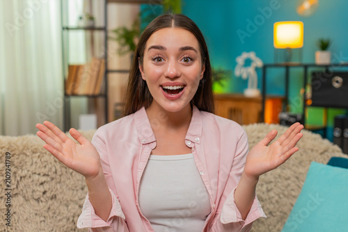 Young woman sits on home sofa reading smartphone then shouts wow eyes wide with joyful surprise at news. Caucasian girl beams happily astonished by unexpected positive message and lifts hand excitedly