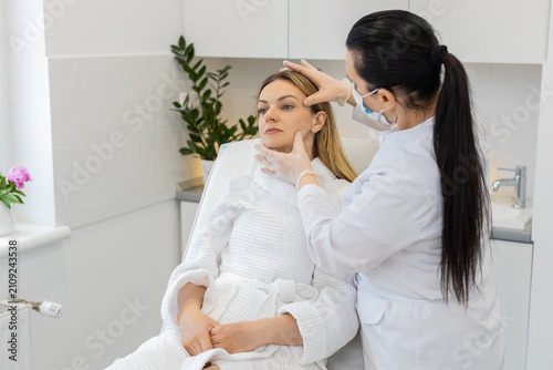 Middle-aged woman sits in chair while cosmetologist in gloves gently checks facial skin before treatment. Calm adult client in white robe enjoys skincare consultation inside modern beauty clinic room.