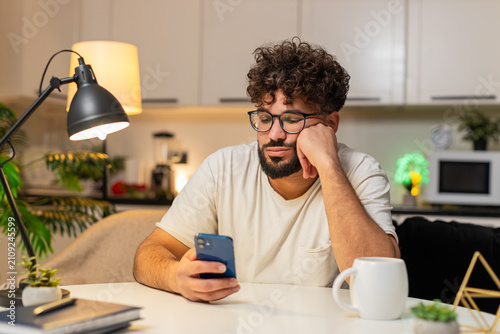 Bild auf Leinwand Arabian man at home scrolls smartphone news feels bored stressed, negative expression, sighs wants rest