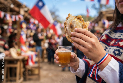 Hand holding a Chilean empanada and a drink during Fiestas Patrias celebration with flags in the background. Independence Day.