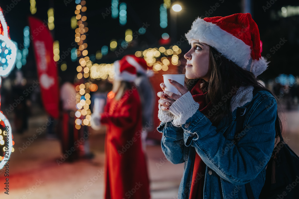 Fototapeta premium Two girls wearing Santa hats enjoy warm drinks at a festive outdoor night market. They stand among holiday lights and decorations, bundled in winter coats.